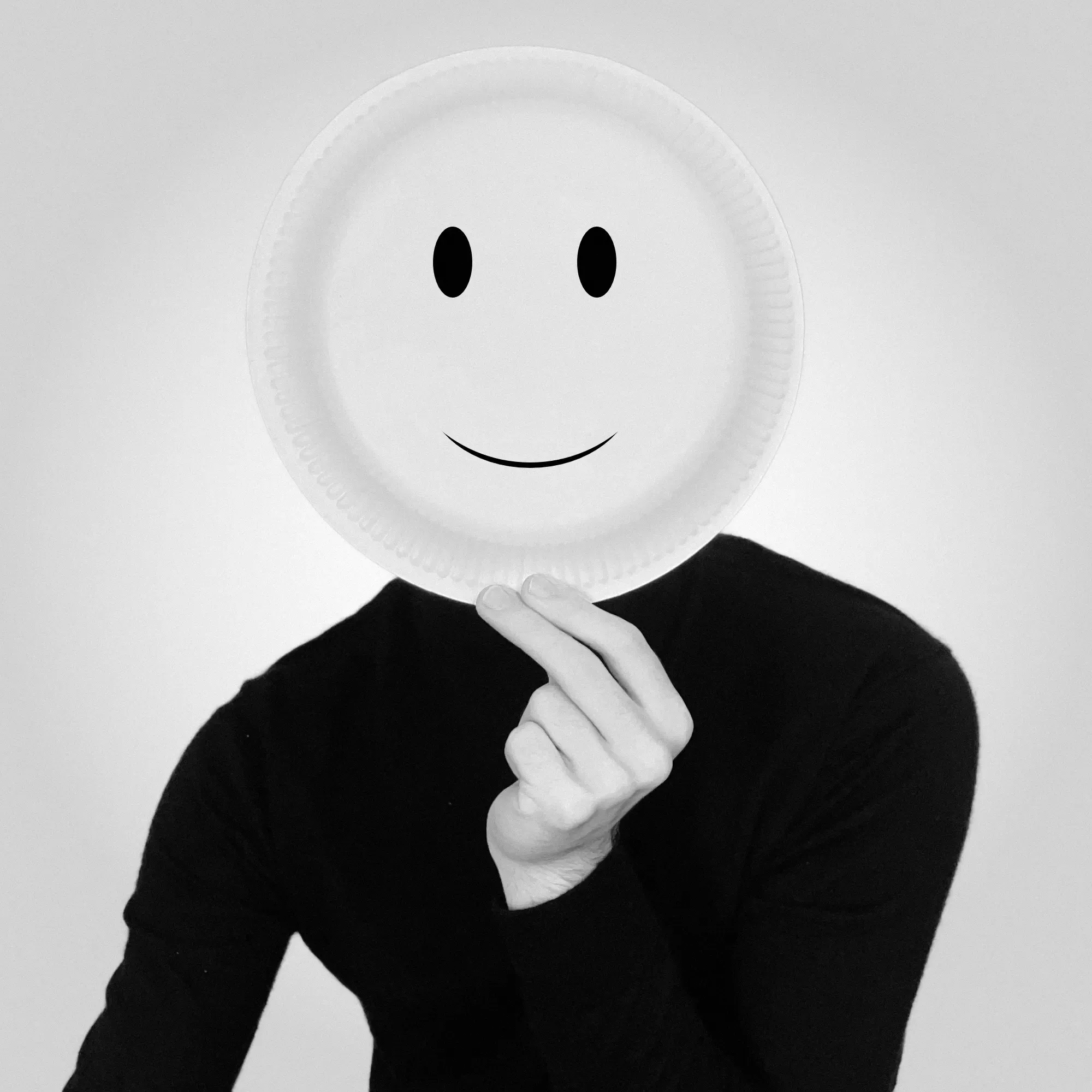 Picture of Jan in grayscale, hiding his face behind a paper plate with a smiley on it.